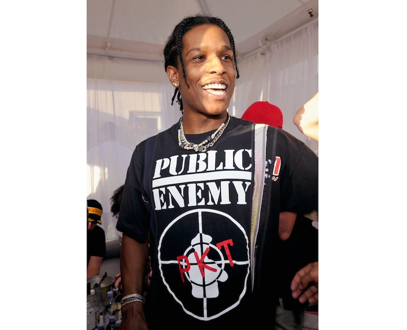 May include: A man wearing a black t-shirt with a white "Public Enemy" logo and a red "P.K.T." logo. He is smiling and wearing a silver chain necklace.