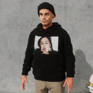 May include: A black hoodie with a graphic print of a woman's face and the word "LOVE" on the front. The person wearing the hoodie is also wearing a black beanie.