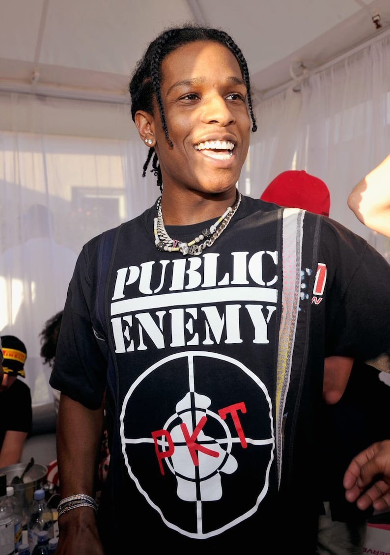 May include: A man wearing a black t-shirt with a white "Public Enemy" logo and a red and white target symbol. The target symbol has the letters "PKT" written in red inside of it.