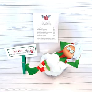 May include: A whimsical spa-themed display featuring an elf figurine relaxing in a clear bowl filled with cotton. A sign reads "Spa Day" and a spa menu lists treatments. The elf wears a white towel and cucumber slices on its eyes.