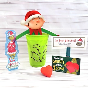 May include: A festive Grinch-themed decoration set. Includes a green cup with a Grinch face, a red heart, and signs that read "I've been Grinched!" and "Grow the GRINCH'S Heart 3 Sizes!" A Grinch character and elf are also included.