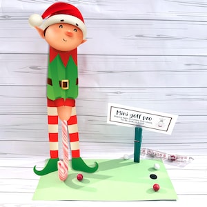 May include: A whimsical Christmas-themed mini golf set featuring an elf figure with a red hat and green outfit. The set includes a green putting green, candy cane golf club, and a sign that reads "Mini golf pro".