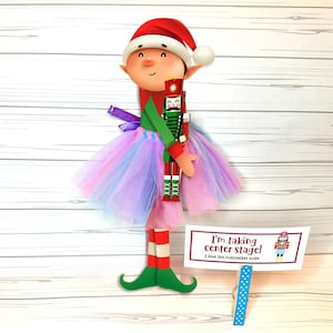 May include: A whimsical Christmas elf figure wearing a red hat, a tutu, and holding a nutcracker. The elf has green shoes and red and white striped legs. A sign reads "I'm taking center stage! I love the nutcracker suite."