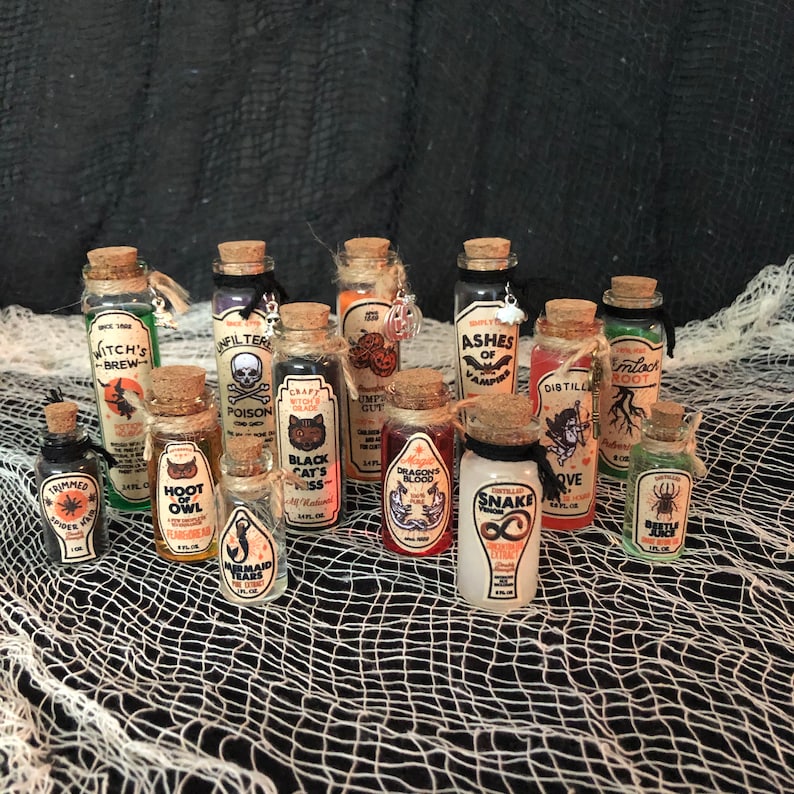 Op de afbeelding: Een verzameling van 14 kleine glazen flesjes met kurkstoppen en Halloween-thema labels. De labels bevatten verschillende griezelige en fantasierijke ontwerpen, waaronder heksen, uilen, slangen en draken. De flesjes zijn gevuld met kleurrijke vloeistoffen en zijn gerangschikt op een zwart en wit net.