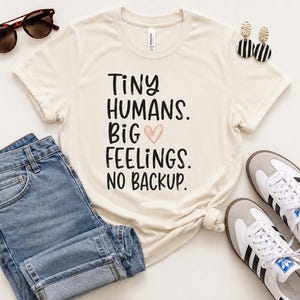 Può includere: T-shirt color crema con la scritta "Tiny Humans. Big Feelings. No Backup." in nero e un cuore rosa. La maglietta è abbinata a jeans blu, occhiali da sole, orecchini e scarpe da ginnastica.