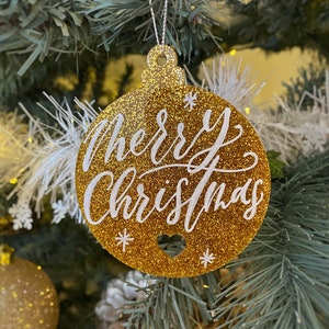 May include: A round, gold glitter Christmas ornament featuring the white script text Merry Christmas. The ornament has a small heart-shaped cutout at the bottom and is suspended by a thin silver string, displaying white star accents on the surface.