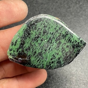 May include: A green and black gemstone with a mottled pattern. The stone is irregularly shaped and has a smooth, polished surface.