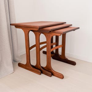 May include: A set of three wooden nesting tables in a dark brown finish. Each table has a rectangular top and distinctive arched legs. The tables are arranged in a nested formation, with the smallest table fitting under the larger ones.