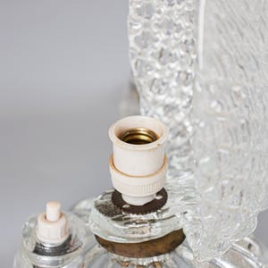 May include: A clear glass lamp base featuring a textured, rippled surface design. The lamp includes a central white socket with a brass-colored interior, a small white push-button switch, and a metallic base ring connecting the glass components.