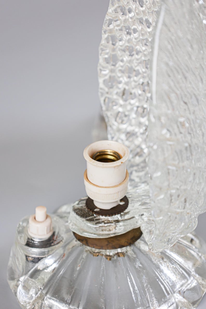 May include: A clear glass lamp base featuring a textured, rippled surface design. The lamp includes a central white socket with a brass-colored interior, a small white push-button switch, and a metallic base ring connecting the glass components.