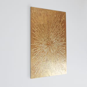 Op de afbeelding: Een rechthoekig, goudkleurig wandkunstwerk met een getextureerd oppervlak. Het kunstwerk heeft een centraal burst-ontwerp met uitstralende lijnen en kleine cirkelvormige details, waardoor een steruitbarstingseffect ontstaat. Het stuk is op een witte muur gemonteerd.