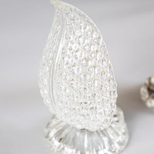 May include: A clear glass decorative object shaped like a leaf or flame with a textured, diamond-patterned surface. The object features a pointed top and rests on a circular, fluted base, reflecting light across its multifaceted, transparent exterior.