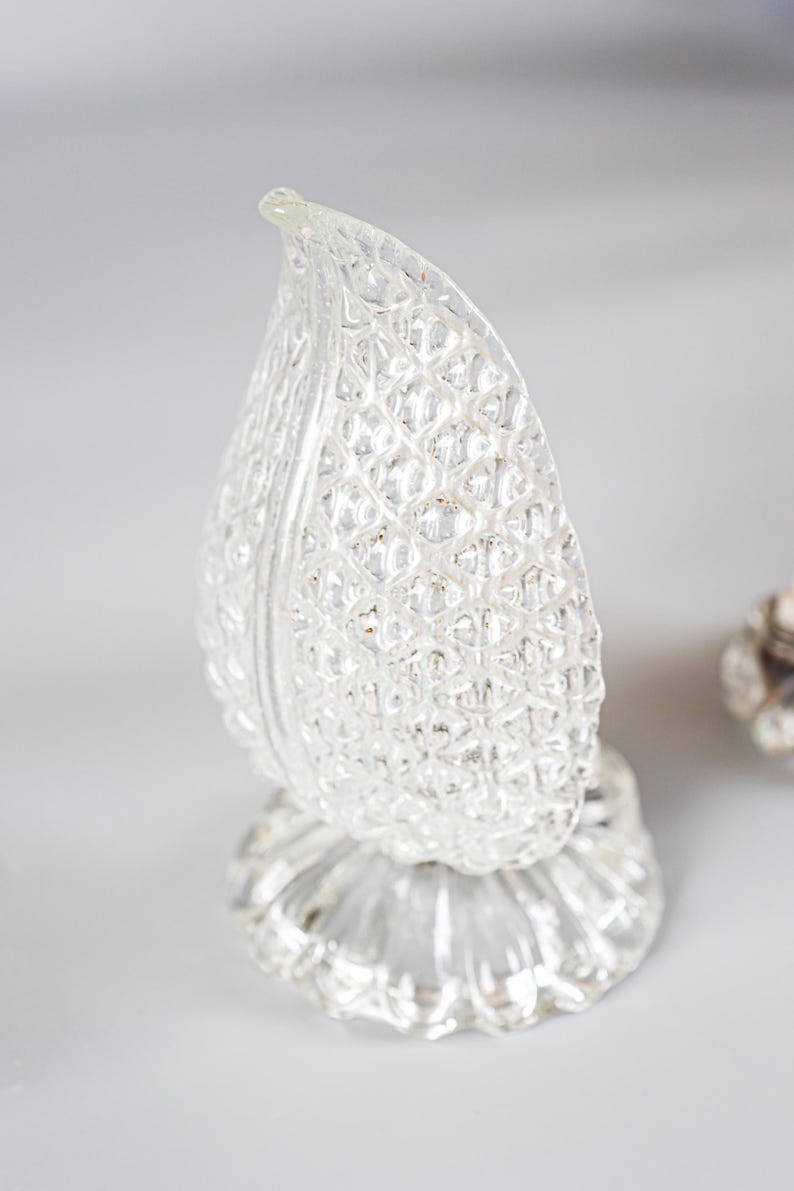 May include: A clear glass decorative object shaped like a leaf or flame with a textured, diamond-patterned surface. The object features a pointed top and rests on a circular, fluted base, reflecting light across its multifaceted, transparent exterior.