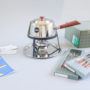 Op de afbeelding: Rvs fonduepan met een houten handvat en deksel, op een zwarte metalen standaard en een zilveren dienblad. Inclusief dozen met fonduevorken en een blauwe brochure met gele afbeeldingen. De pan is voorzien van de naam "Alessi".
