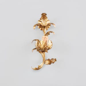 Peut inclure: Un appliques mural dorée avec un motif floral. L'applique présente des feuilles ornées et stylisées et contient une seule bougie. La bougie est de couleur pêche clair. L'applique est fixée sur un mur gris clair.