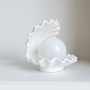 Op de afbeelding: Een witte keramische lamp in de vorm van een open schelp met een gladde, bolvormige witte glazen bol aan de binnenkant. De schelp heeft een geschulpte rand en gestructureerde ribbels op het binnenoppervlak, wat zorgt voor een heldere uitstraling.