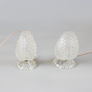 May include: A pair of clear glass lamps with a textured, pineapple-like pattern on the exterior. Each lamp features a rounded, bulbous shade with a vertical seam and a fluted, circular base. A thin, light-colored electrical cord extends from each.