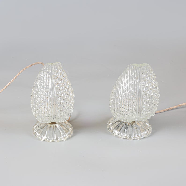 May include: A pair of clear glass lamps with a textured, pineapple-like pattern on the exterior. Each lamp features a rounded, bulbous shade with a vertical seam and a fluted, circular base. A thin, light-colored electrical cord extends from each.