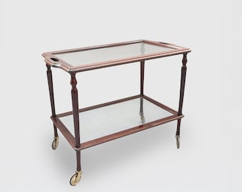 May include: A two-tiered mahogany serving cart with glass shelves and brass wheels. The cart has a rectangular shape and is on a white background.