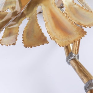 Op de afbeelding: Een gouden metalen sculptuur van een blad met een getextureerd oppervlak en een parelrand. Het blad is bevestigd aan een gouden metalen stengel met zilveren accenten.