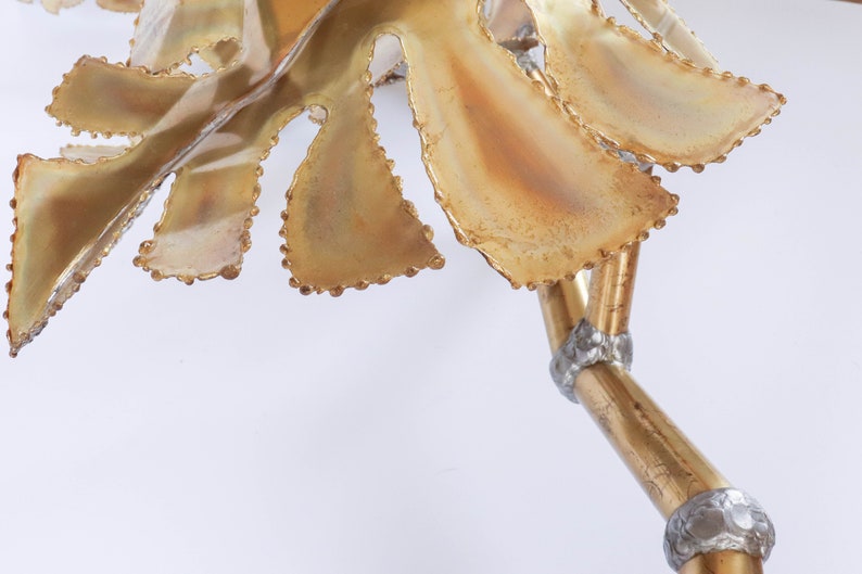 Op de afbeelding: Een gouden metalen sculptuur van een blad met een getextureerd oppervlak en een parelrand. Het blad is bevestigd aan een gouden metalen stengel met zilveren accenten.