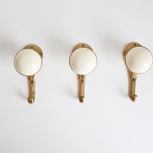 May include: Three brass-coloured wall hooks with ivory-coloured, rounded knobs. The hooks are arranged horizontally against a white background. The hooks have a classic, vintage design, suitable for hanging coats, towels, or decorative items.