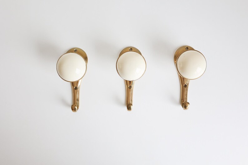 May include: Three brass-coloured wall hooks with ivory-coloured, rounded knobs. The hooks are arranged horizontally against a white background. The hooks have a classic, vintage design, suitable for hanging coats, towels, or decorative items.