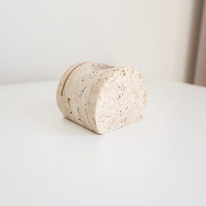 May include: A beige, cylindrical, stone-like object with a slot cut into the top. The object has a rough, textured surface and is positioned on a white surface. The object appears to be a decorative item.