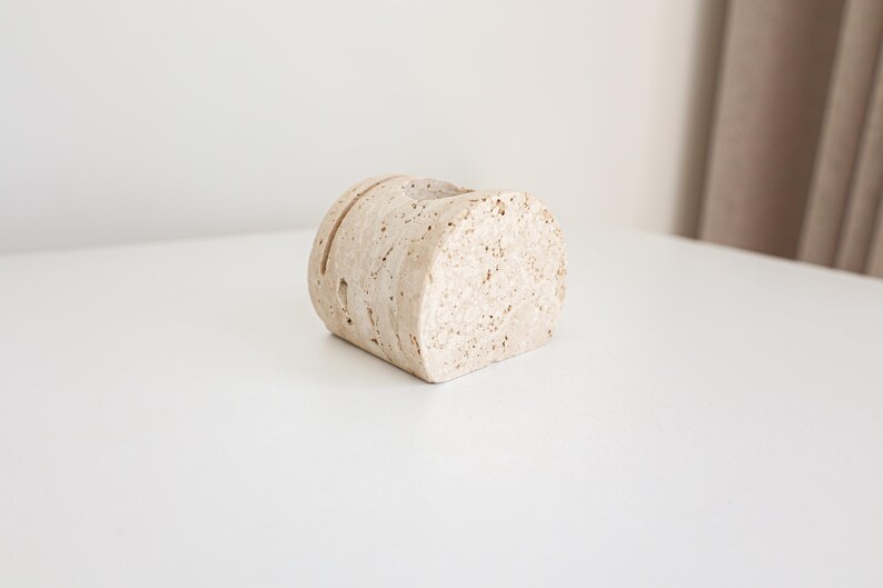 May include: A beige, cylindrical, stone-like object with a slot cut into the top. The object has a rough, textured surface and is positioned on a white surface. The object appears to be a decorative item.