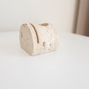 May include: A beige, rectangular desk organizer made of stone. The organizer has a slot on top and a small hole. The stone has a textured surface with small holes throughout. The organizer is on a white surface.
