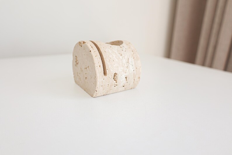 May include: A beige, rectangular desk organizer made of stone. The organizer has a slot on top and a small hole. The stone has a textured surface with small holes throughout. The organizer is on a white surface.