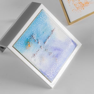 May include: Two square abstract art pieces. One is angled, featuring a white frame and a blue, purple, and orange design. The second piece, with a gold frame, displays an orange splatter design on a white background. Both pieces are on a white surface.