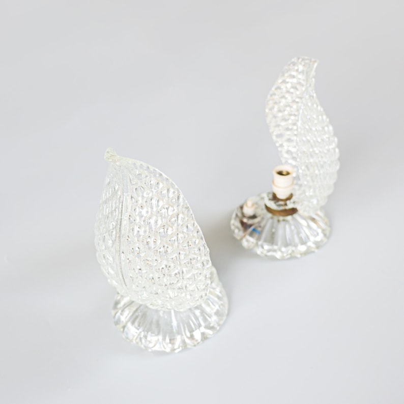 May include: Two clear glass table lamps featuring a textured, leaf-shaped shade with a diamond pattern. Each lamp has a circular, fluted base made of clear glass. One lamp is fully assembled, while the other shows the internal light bulb socket.