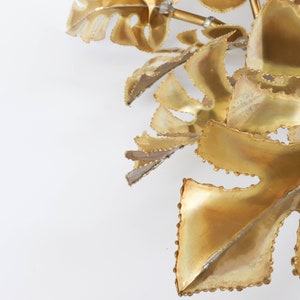 Op de afbeelding: Een gouden metalen wandsculptuur met een cluster van gestileerde bladeren met een textuur.