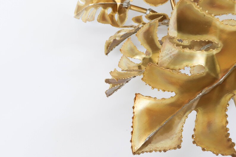 Op de afbeelding: Een gouden metalen wandsculptuur met een cluster van gestileerde bladeren met een textuur.
