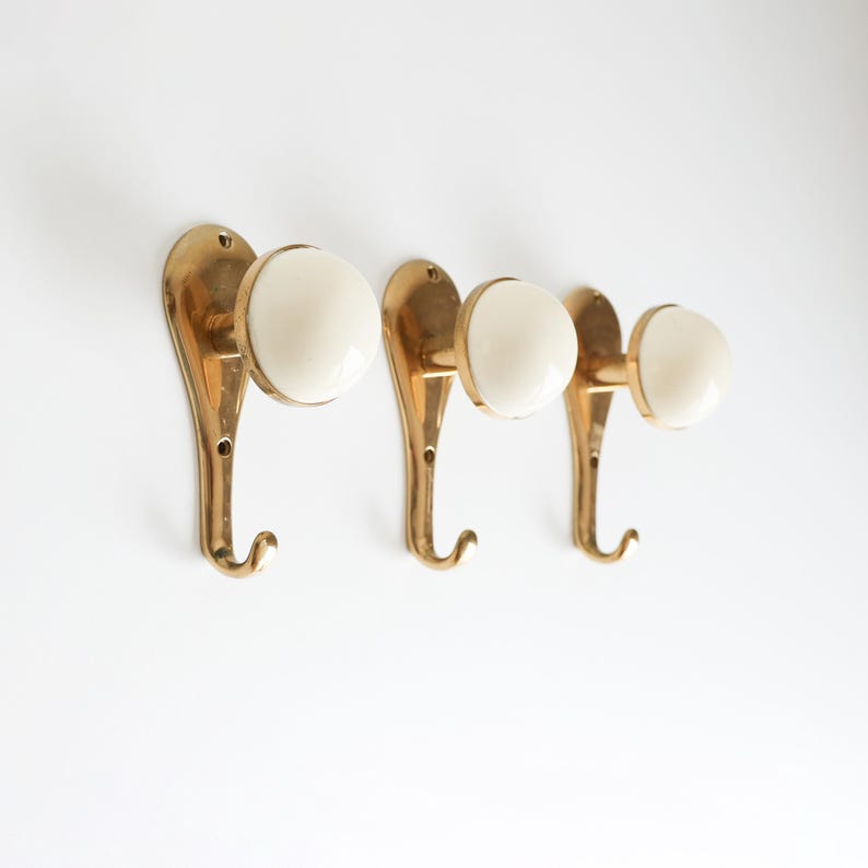 May include: Three brass wall hooks with cream ceramic knobs. The hooks have a curved design and are mounted on a white background. These decorative hooks are suitable for hanging coats, hats, or other items.
