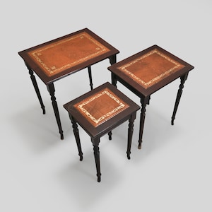 May include: Set of three nesting tables with brown leather tops and decorative gold trim. The tables have turned legs and are made of dark wood.