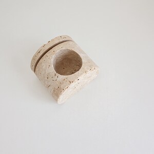 May include: A beige, stone-like candle holder with a circular opening and a groove on top. The holder has a textured surface and is set against a white background. The design is minimalist and modern.