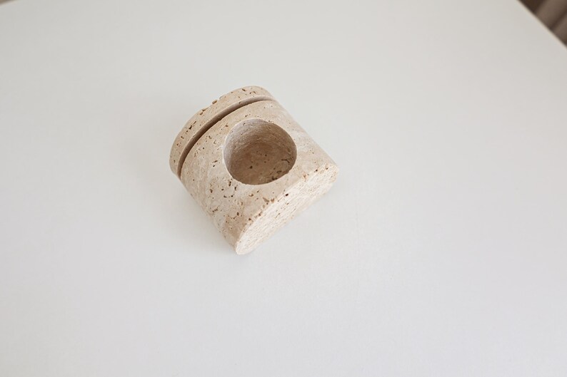 May include: A beige, stone-like candle holder with a circular opening and a groove on top. The holder has a textured surface and is set against a white background. The design is minimalist and modern.