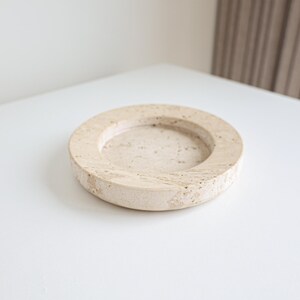 Op de afbeelding: Een beige, cirkelvormige stenen onderzetter met een verzonken midden. De onderzetter is gemaakt van een lichtgekleurde steen met een textuur. De onderzetter staat op een wit oppervlak.