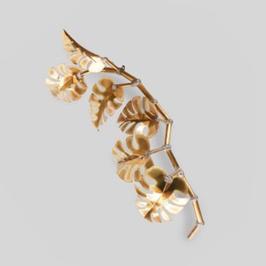 Op de afbeelding: Een gouden metalen tak met vijf bladeren, elk blad is gevormd als een monstera-blad. De bladeren zijn verbonden met de tak met kleine gouden metalen connectoren. De tak is gebogen en heeft een kleine gouden metalen bol aan het einde.