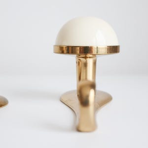 May include: Two brass-coloured wall hooks with a cream-coloured, dome-shaped top. The hooks have a rounded base and a curved hook design. The hooks are displayed against a white background.