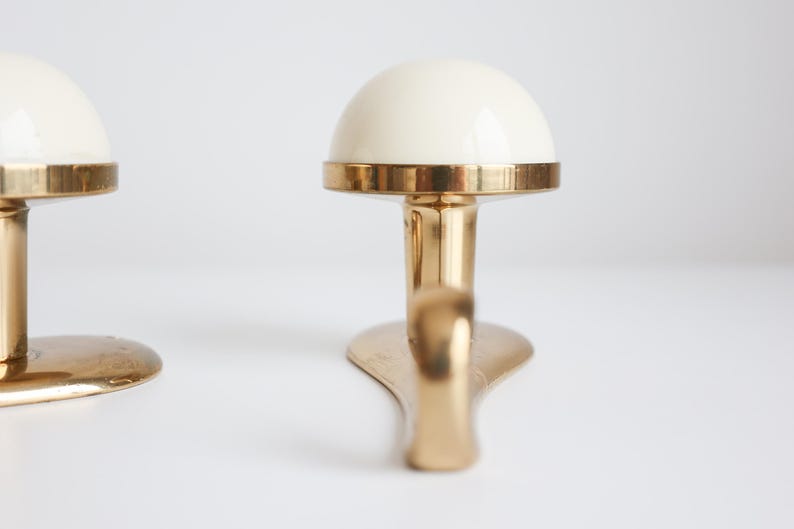 May include: Two brass-coloured wall hooks with a cream-coloured, dome-shaped top. The hooks have a rounded base and a curved hook design. The hooks are displayed against a white background.
