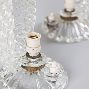 May include: Two clear glass table lamps featuring textured, fan-shaped glass shades and circular, fluted glass bases. Each lamp includes a central white socket for a bulb, a small white push-button switch, and a brass-colored metal connector.