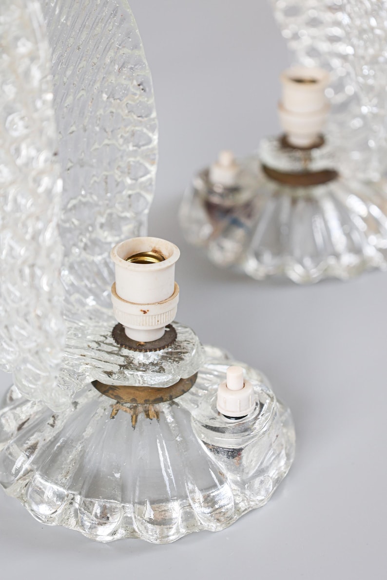May include: Two clear glass table lamps featuring textured, fan-shaped glass shades and circular, fluted glass bases. Each lamp includes a central white socket for a bulb, a small white push-button switch, and a brass-colored metal connector.
