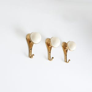 May include: Three vintage brass wall hooks with cream ceramic knobs. The hooks have a curved design and a small hook at the bottom. The hooks are mounted on a white wall.