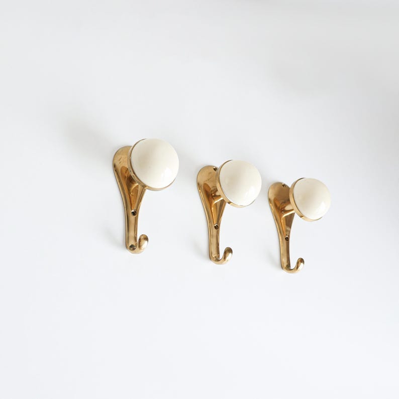 May include: Three vintage brass wall hooks with cream ceramic knobs. The hooks have a curved design and a small hook at the bottom. The hooks are mounted on a white wall.