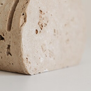 May include: A close-up of a beige, rectangular stone block with a vertical groove. The stone has a rough, textured surface with small holes and imperfections. The object is likely a decorative item or a paper holder.