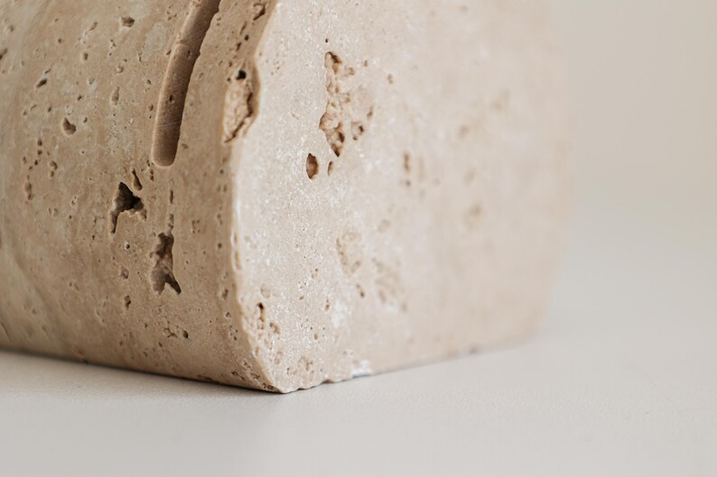 May include: A close-up of a beige, rectangular stone block with a vertical groove. The stone has a rough, textured surface with small holes and imperfections. The object is likely a decorative item or a paper holder.