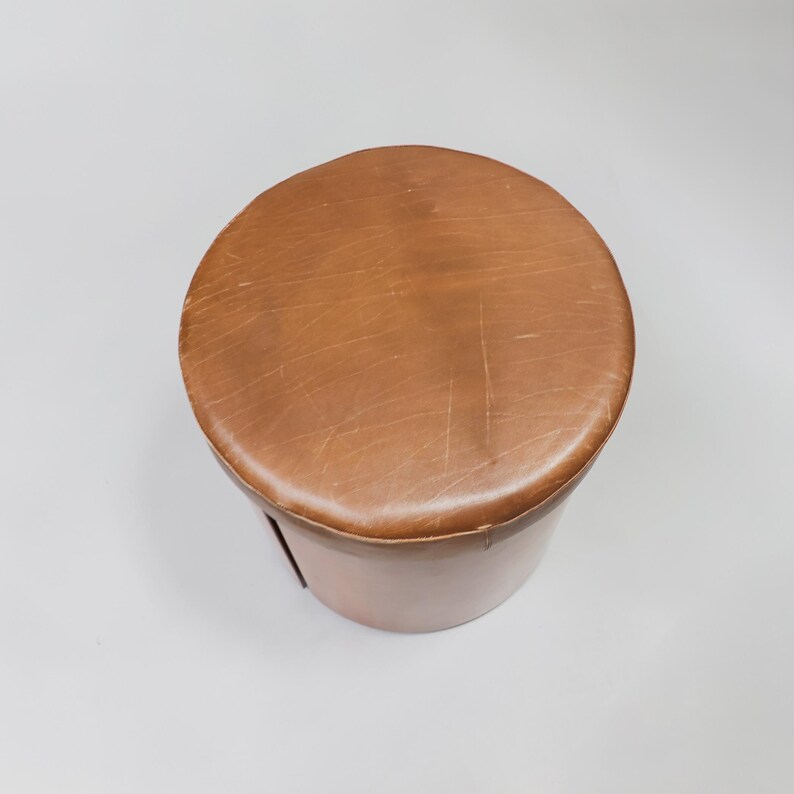 May include: A brown leather ottoman with a round top and a cylindrical base.